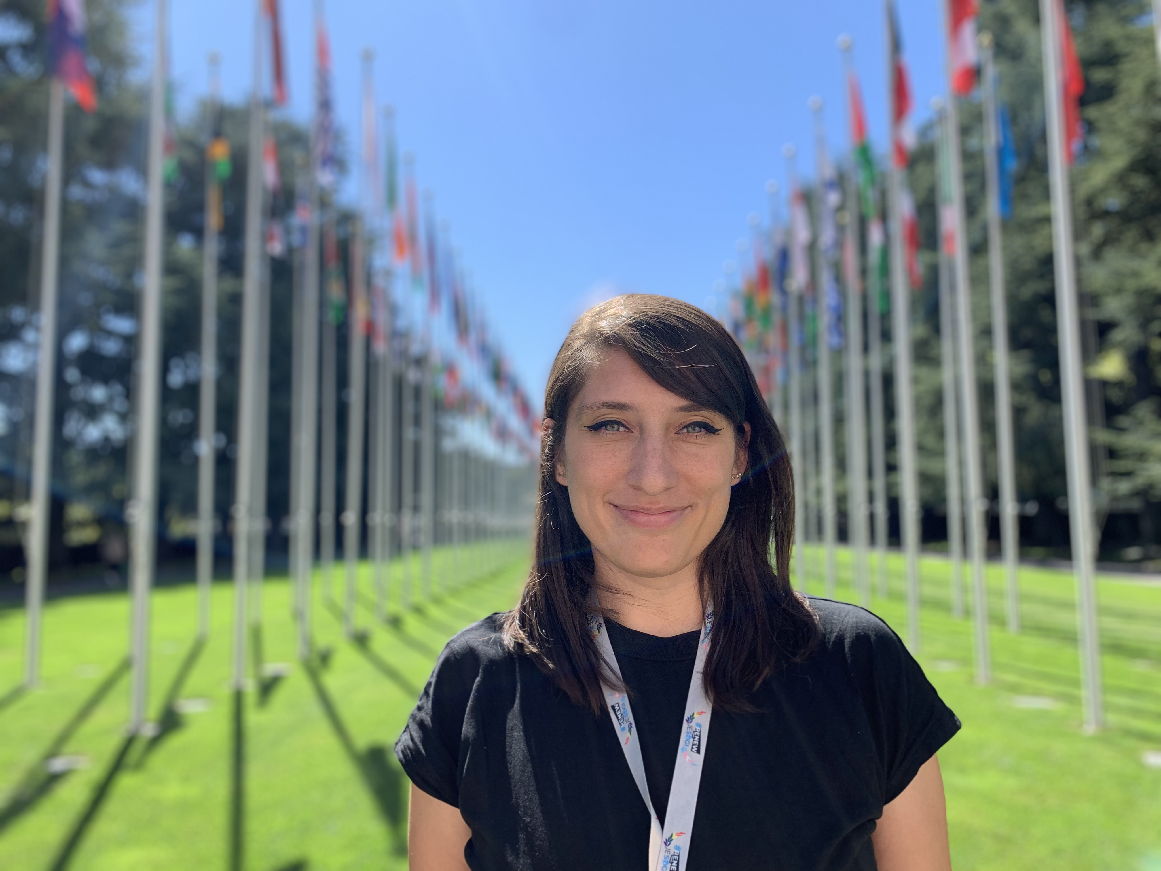 Picture of Paula in a black T-shirt with ID leash on her neck. She has dark brown hair to her collar bones and she's smiling. In the background there are flags of different countries.